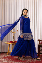 Woman in a blue traditional outfit with silver embroidery standing in a decorated room.