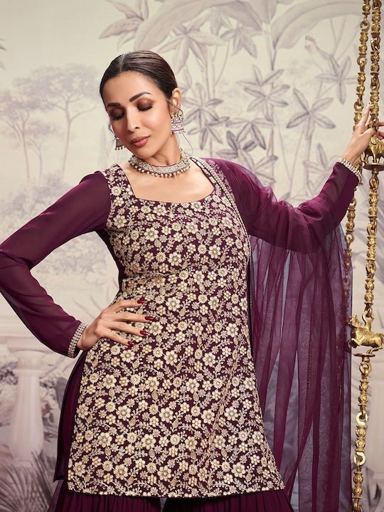 Woman in a floral dress with a purple dupatta against a decorative wall.