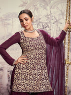 Woman in a floral dress with a purple dupatta against a decorative wall.