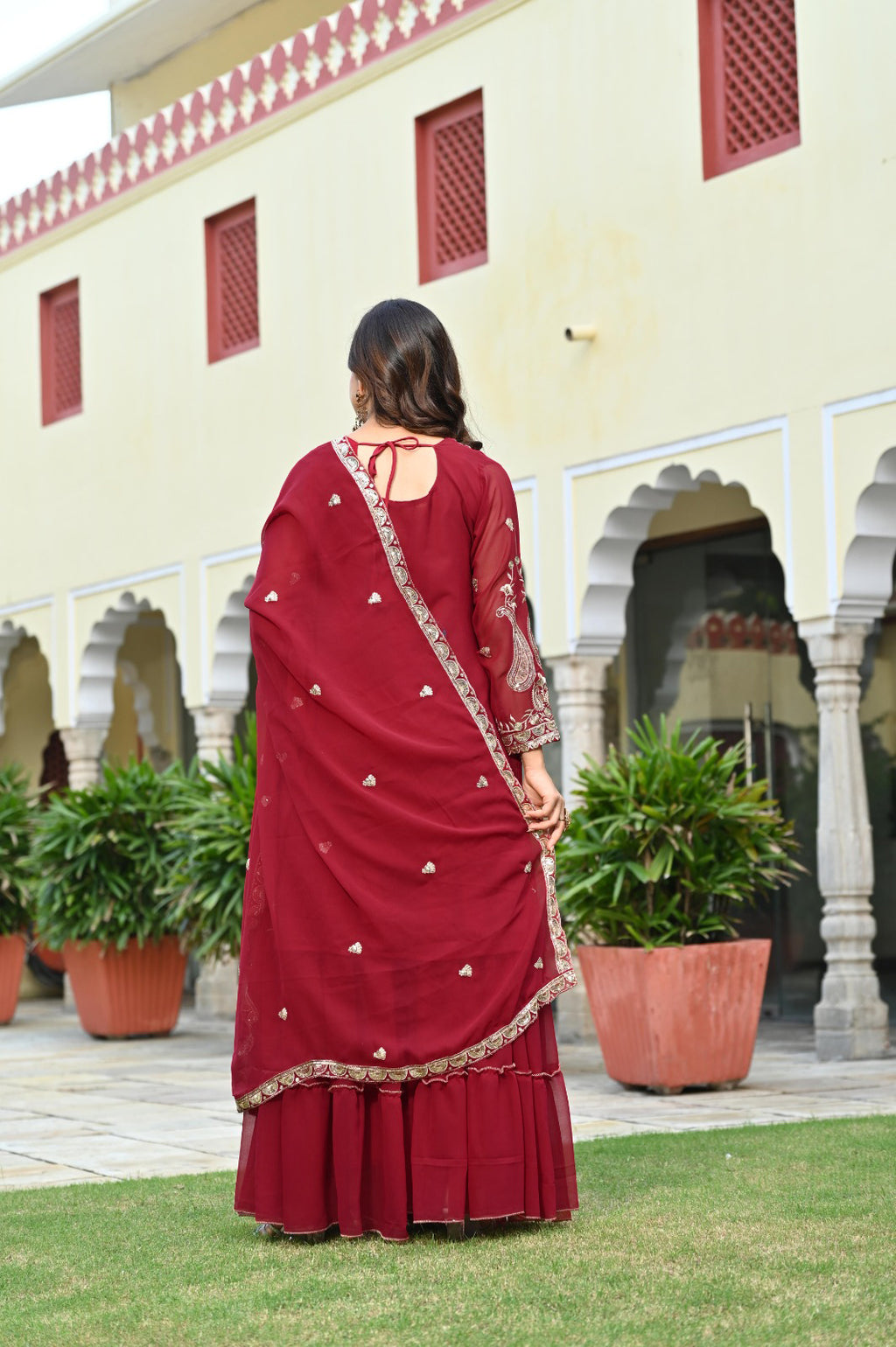 Woman in a maroon dress standing in front of a building with arches and plants.