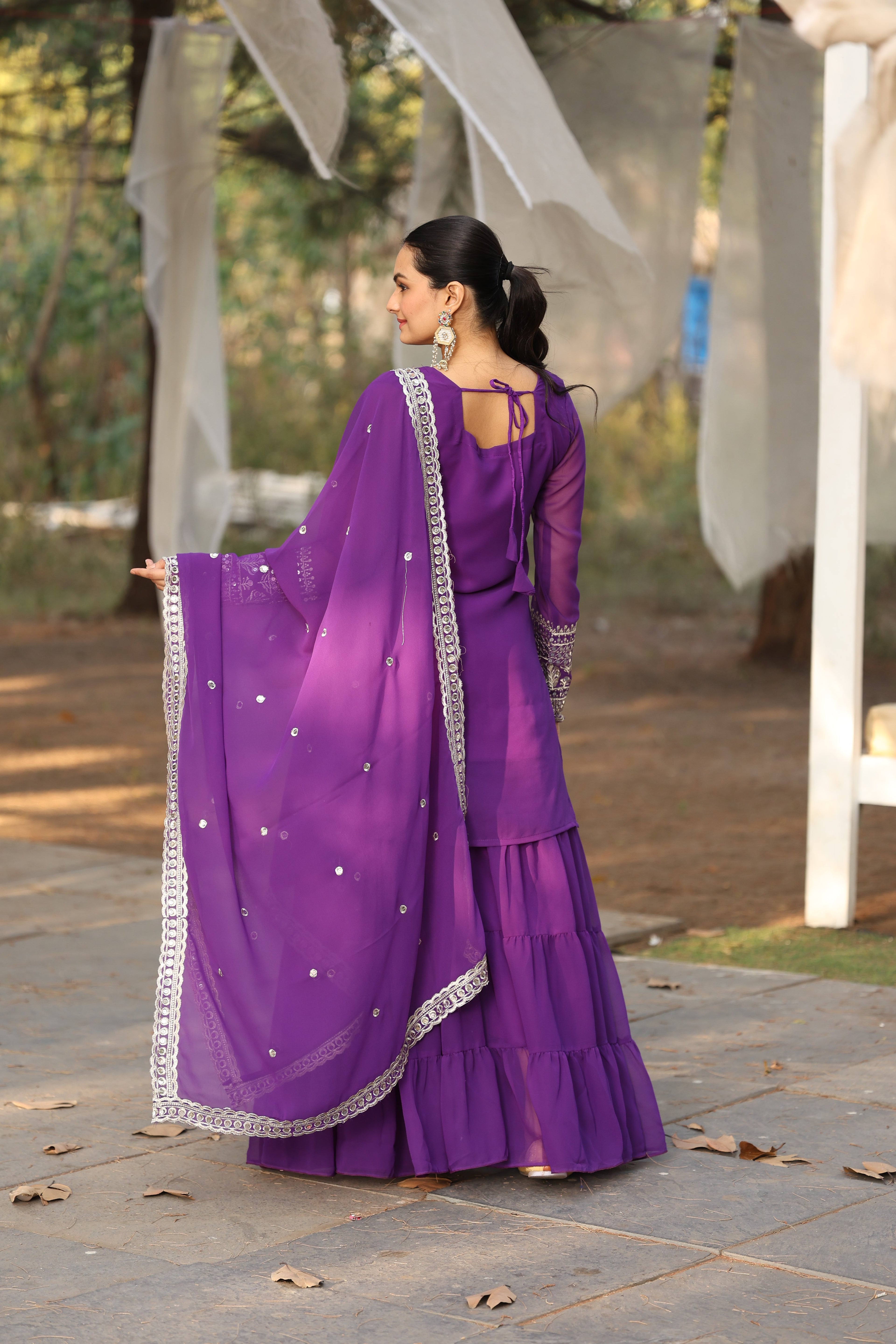 Woman in a purple traditional outfit standing outdoors on a paved path.
