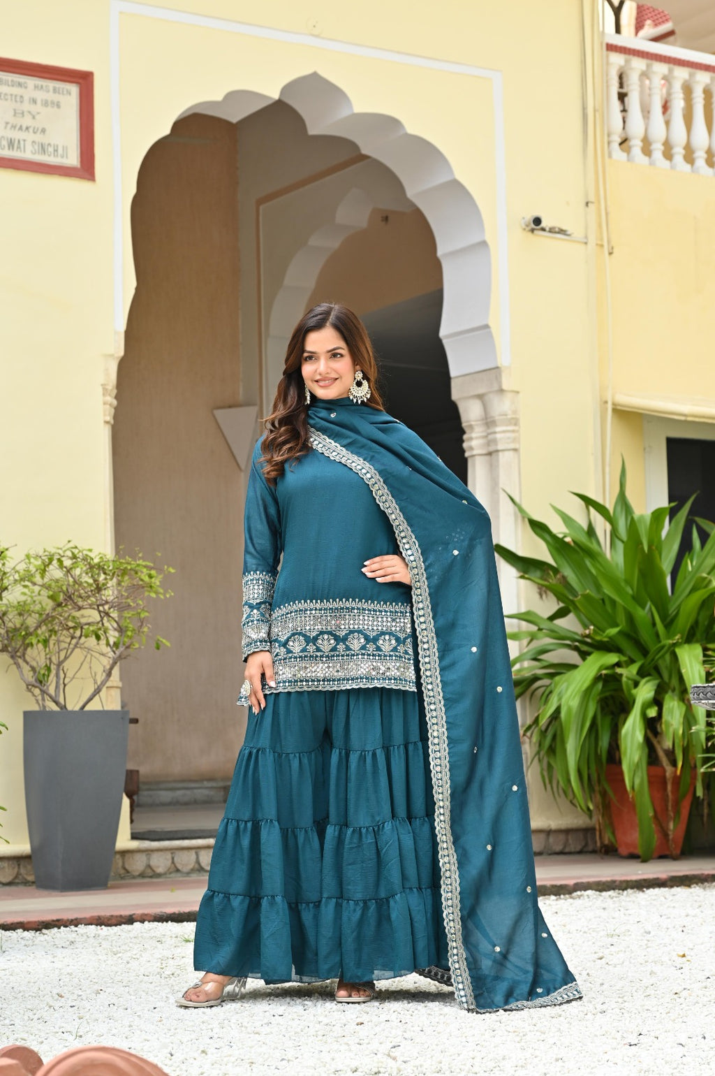 Woman in a teal traditional outfit standing in front of a building with decorative arches.