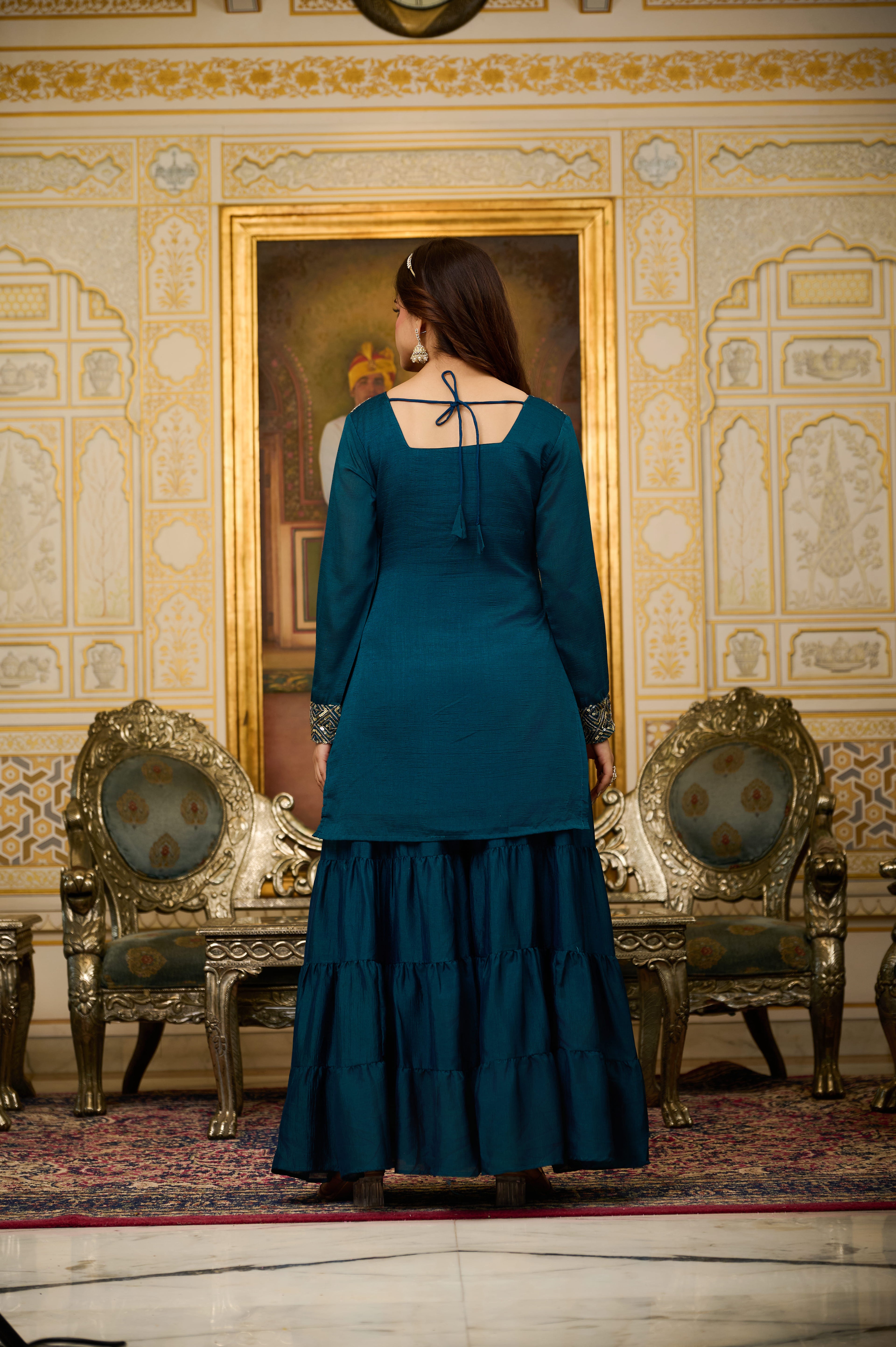 Woman in a teal dress standing in an ornate room with gold accents and a large mirror.