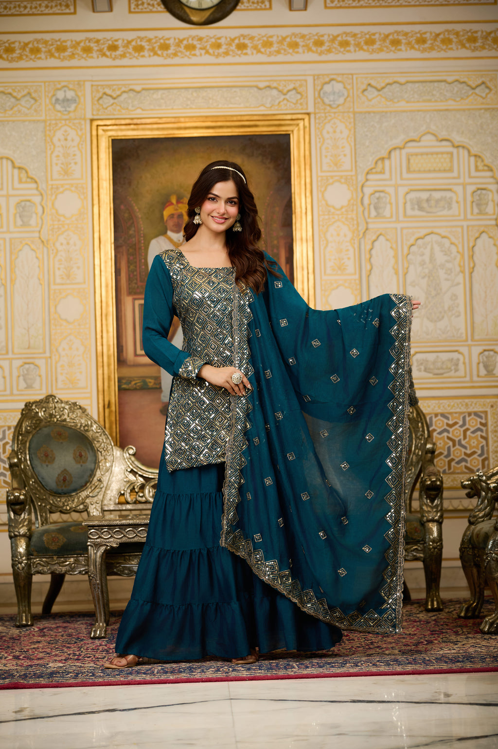 Woman in a blue traditional outfit standing in an ornate room with a painting on the wall.