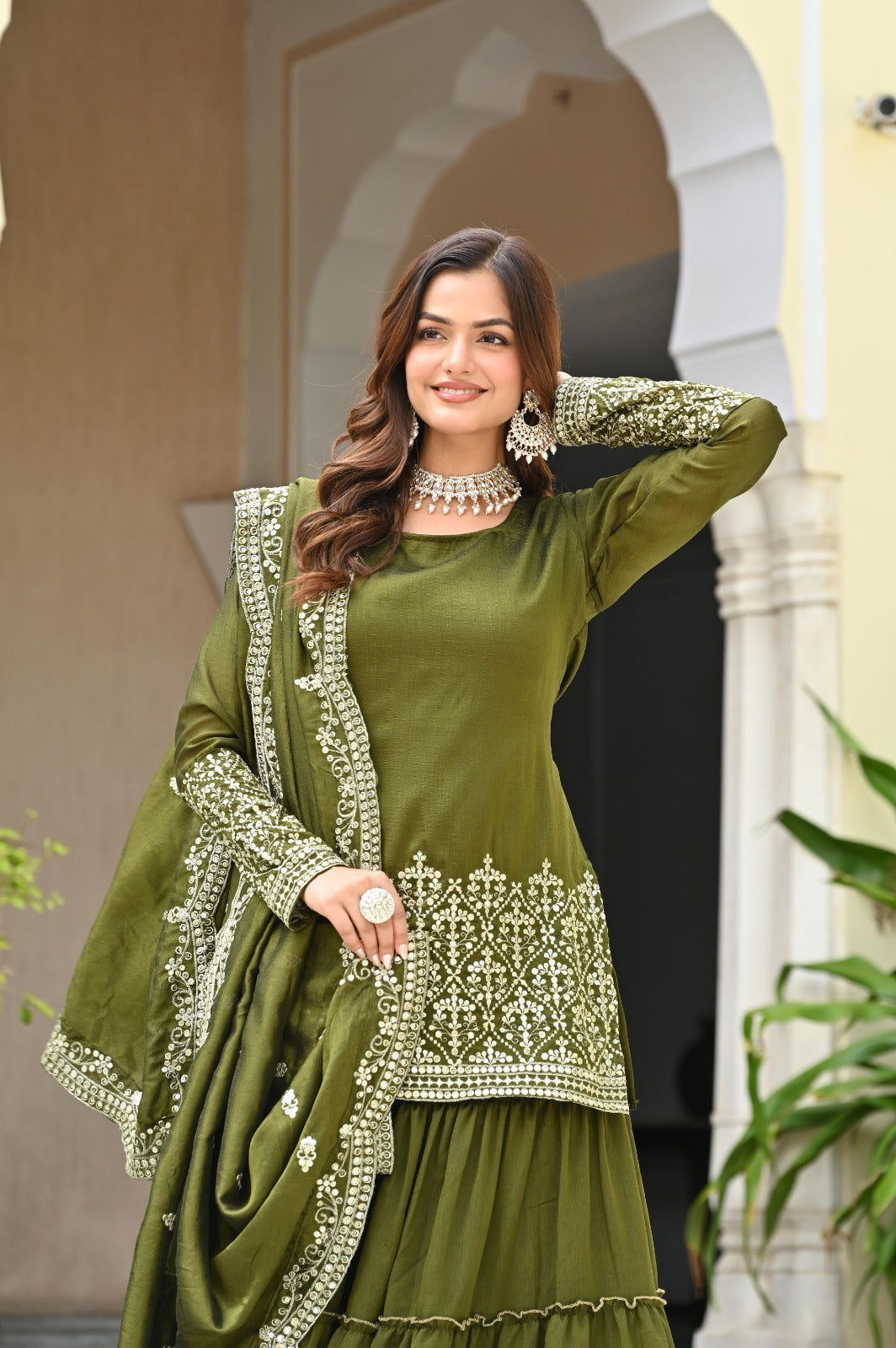 Woman wearing a green traditional outfit with intricate patterns, standing in front of a decorative archway.