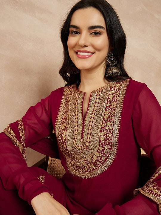 Woman wearing a maroon traditional outfit with gold embroidery on a beige background