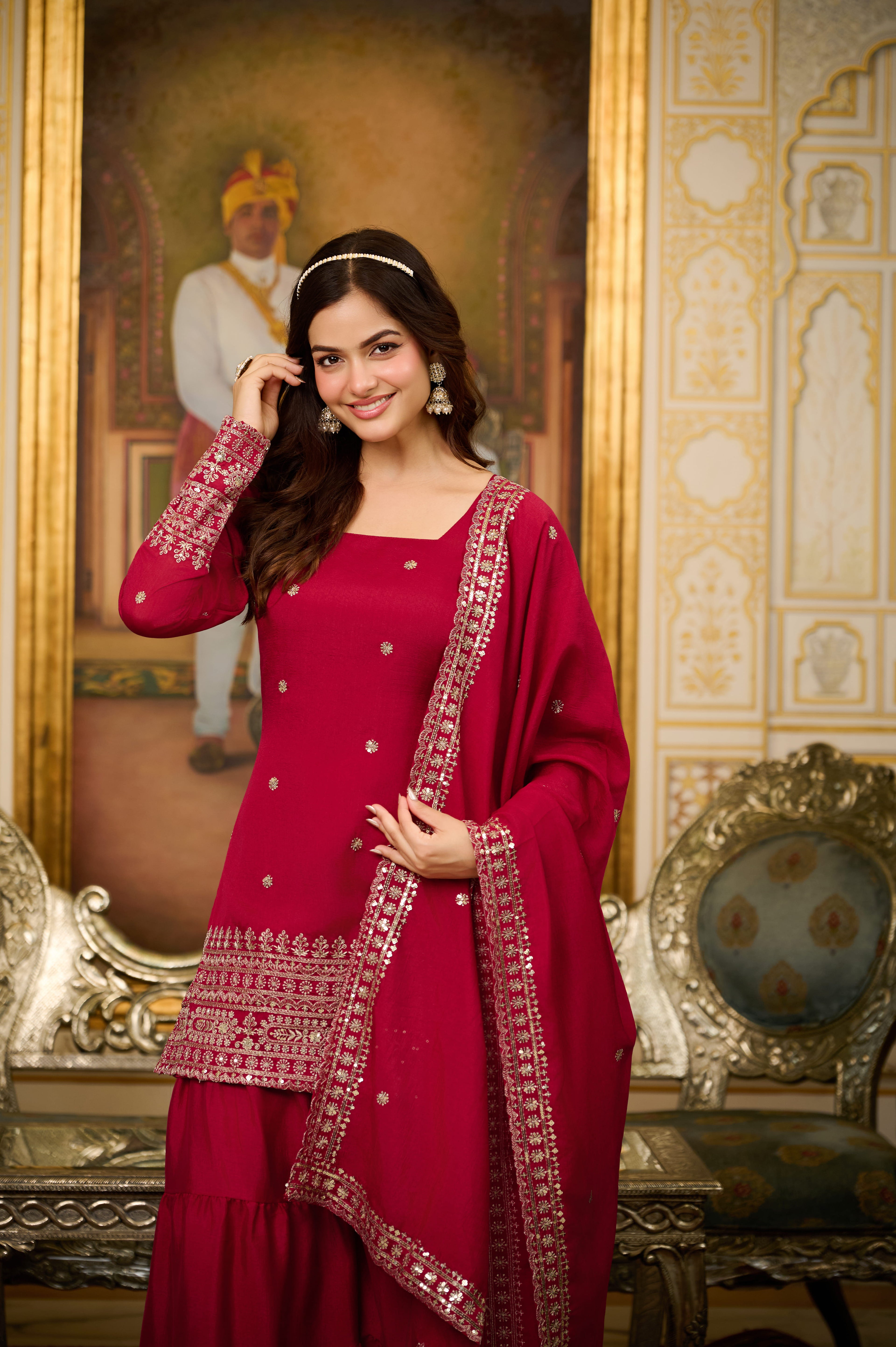 Woman in a red traditional outfit with intricate patterns, standing in an ornate room.