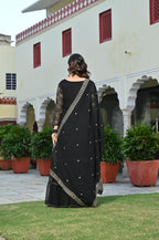 Woman in a black sharara standing in front of a building with red windows and green plants.