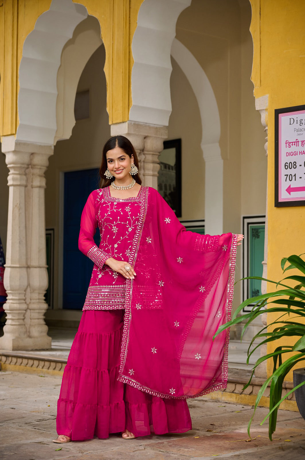 Woman in a bright pink traditional outfit standing in front of a yellow building with arches.