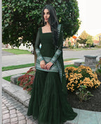 Woman in a dark green traditional outfit standing outdoors with greenery and flowers in the background