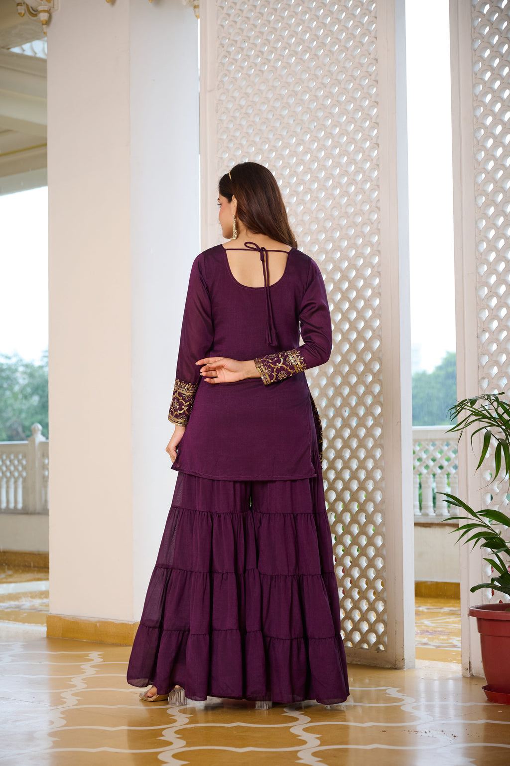 Woman in a purple traditional outfit standing in a well-lit room with white columns and a plant.