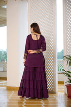 Woman in a purple traditional outfit standing in a well-lit room with white columns and a plant.