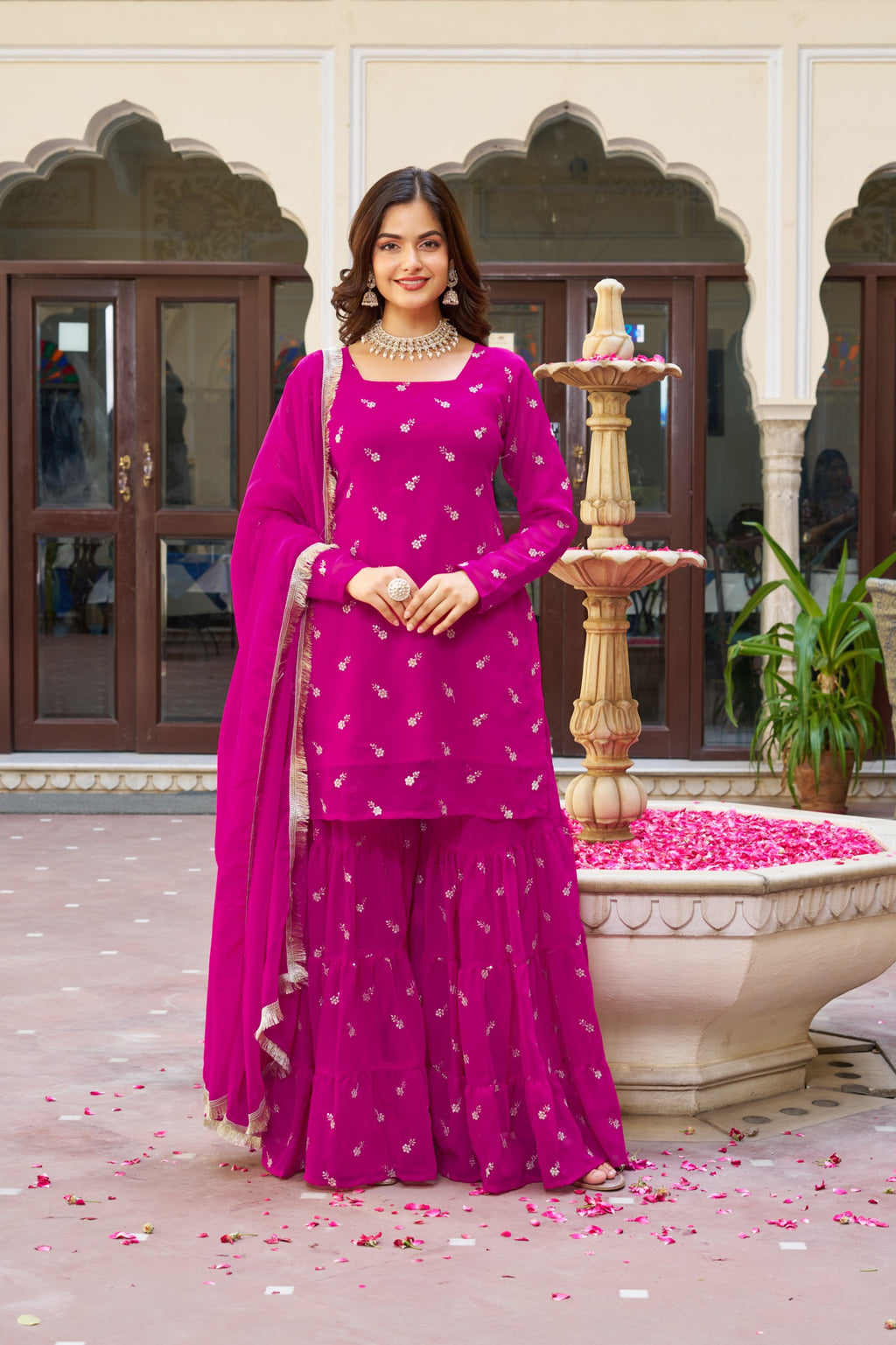 Woman in a pink traditional outfit standing in front of a decorative fountain with pink petals on the ground.