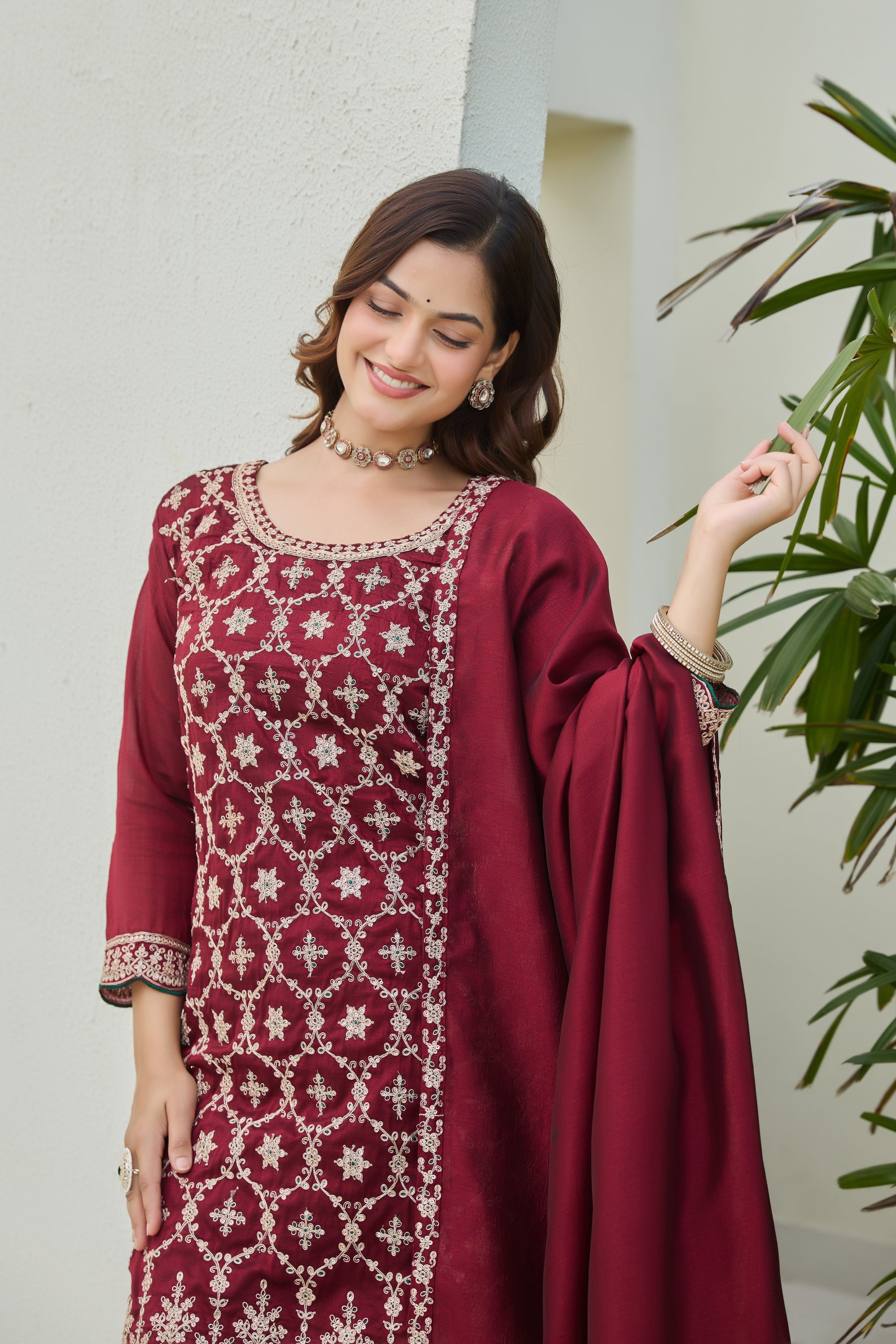 Woman wearing a maroon traditional outfit with white patterns, standing against a light-colored wall with greenery.