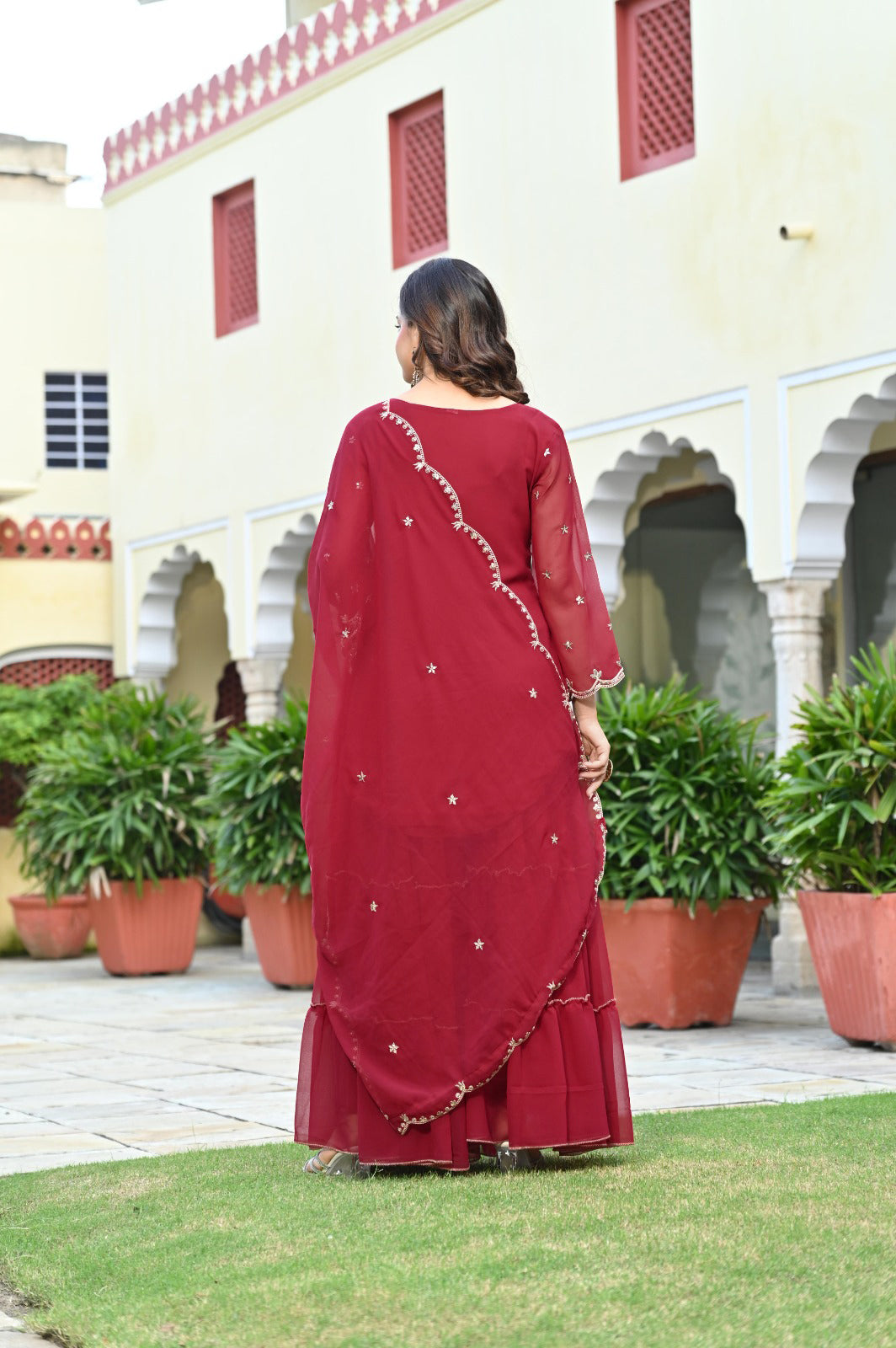 Woman in a maroon traditional outfit standing in front of a building with greenery.
