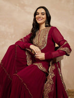 Woman wearing a maroon traditional outfit with gold embroidery against a beige background
