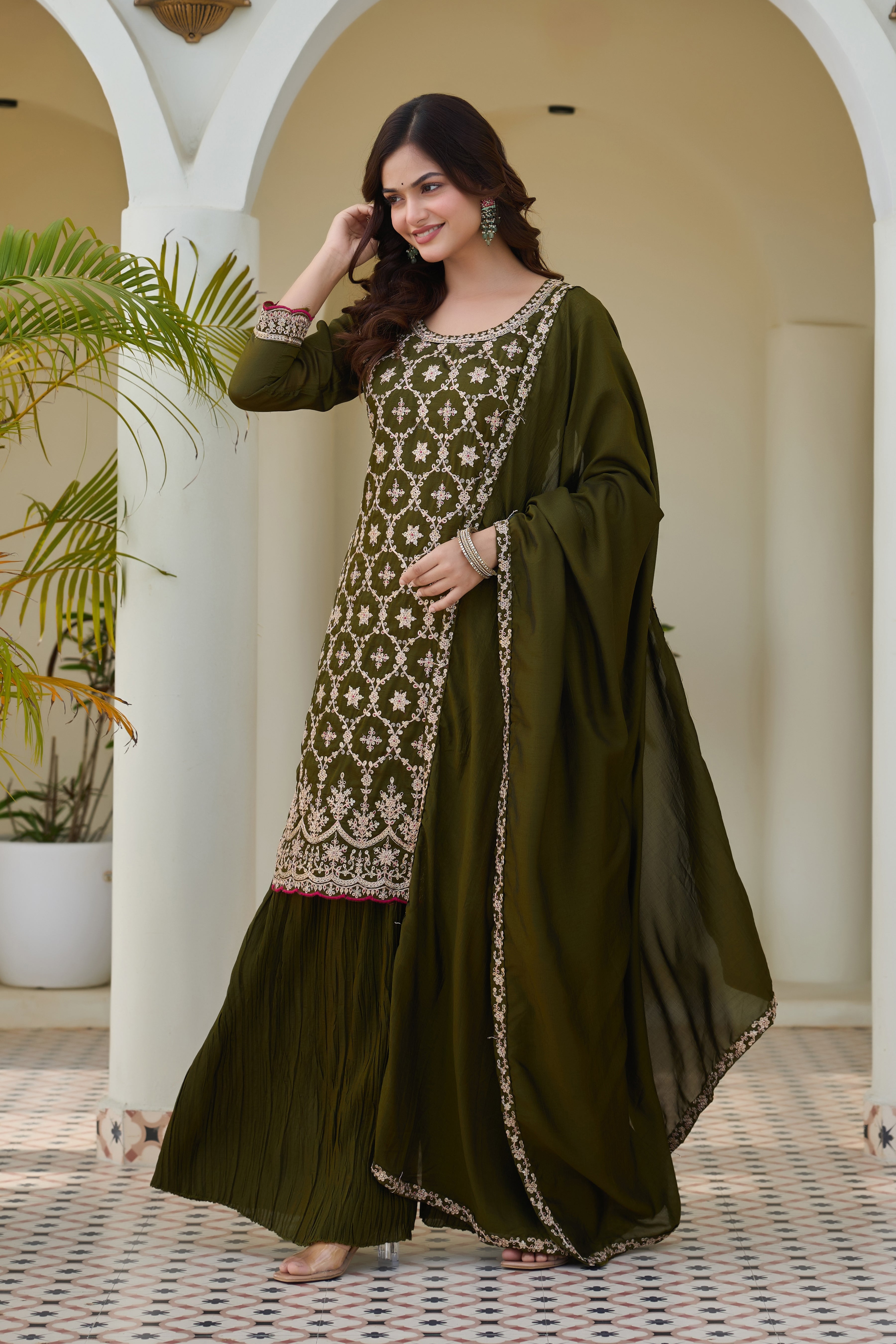 Woman in a green traditional outfit with intricate patterns standing in a decorative indoor setting.