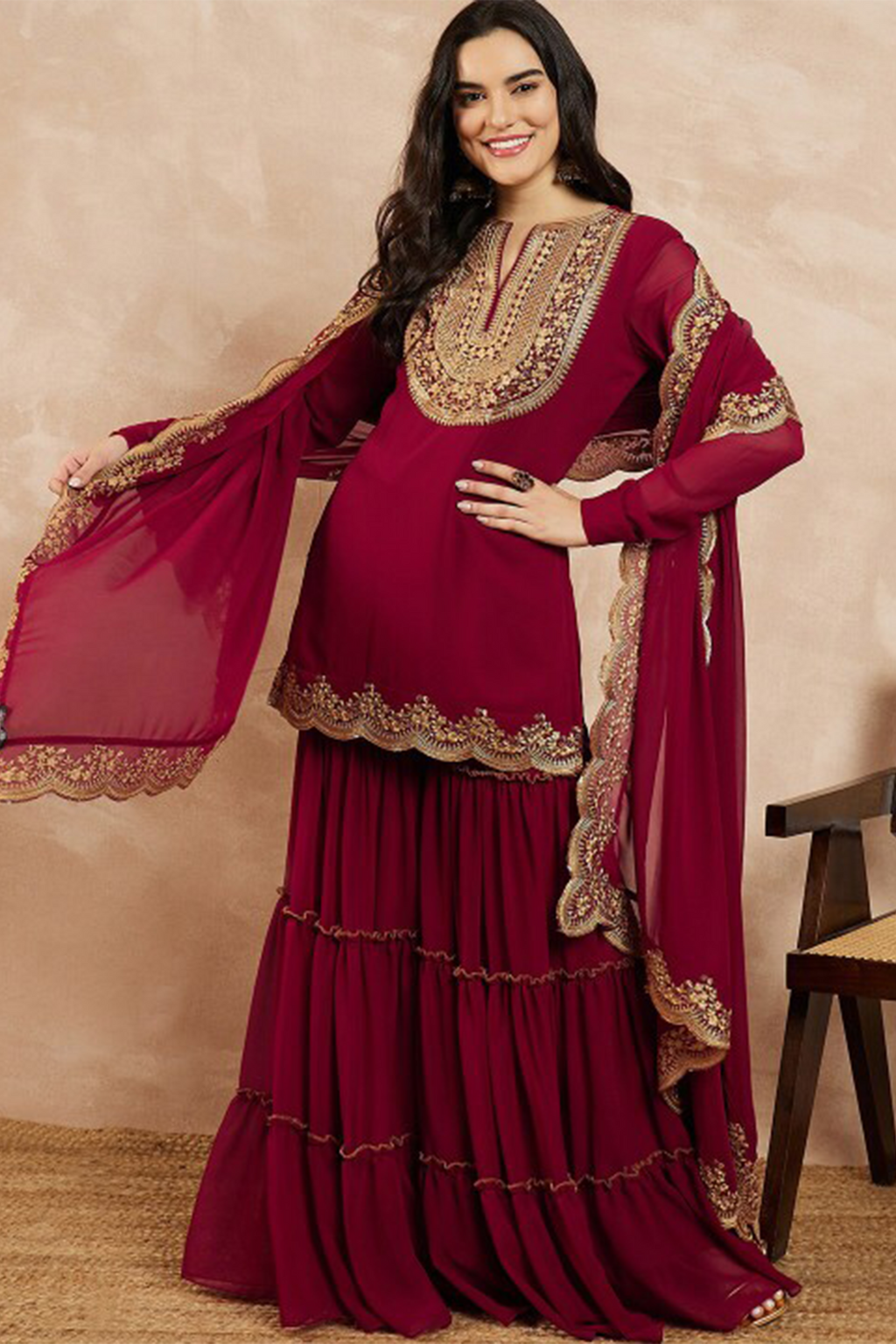 Woman wearing a maroon traditional outfit with gold embroidery against a beige background