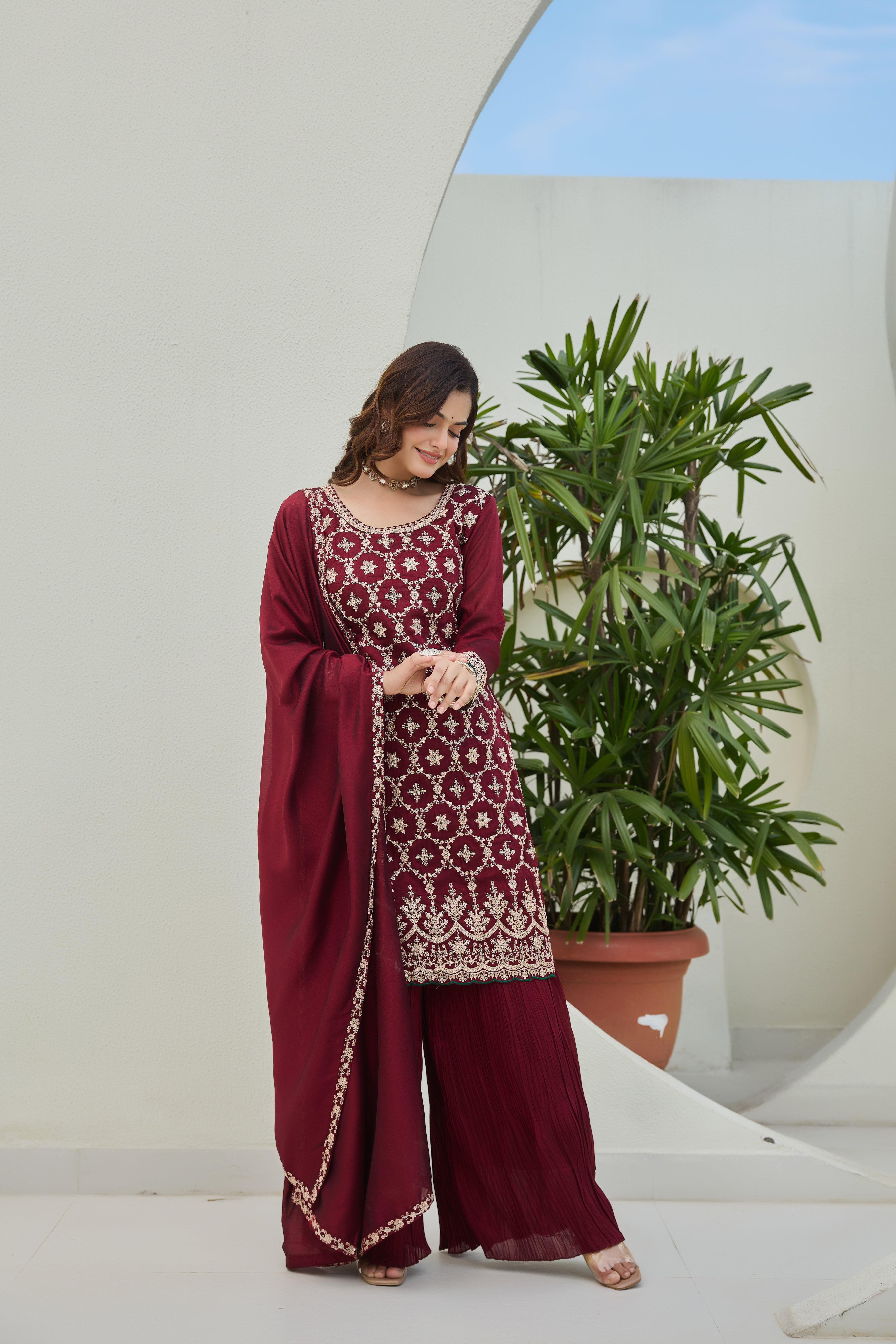 Woman in a maroon traditional outfit standing next to a potted plant indoors.