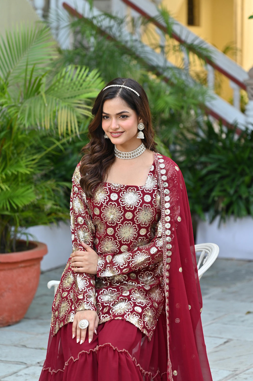 Woman in traditional maroon and gold outfit with jewelry, standing outdoors.