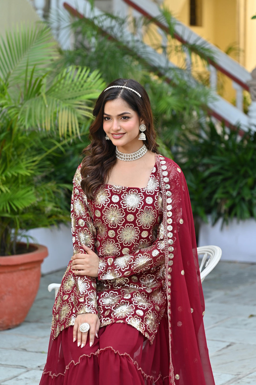 Woman in traditional maroon and gold outfit with jewelry, standing outdoors.