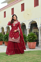 Woman in a maroon traditional outfit standing in front of a building with arches and plants.