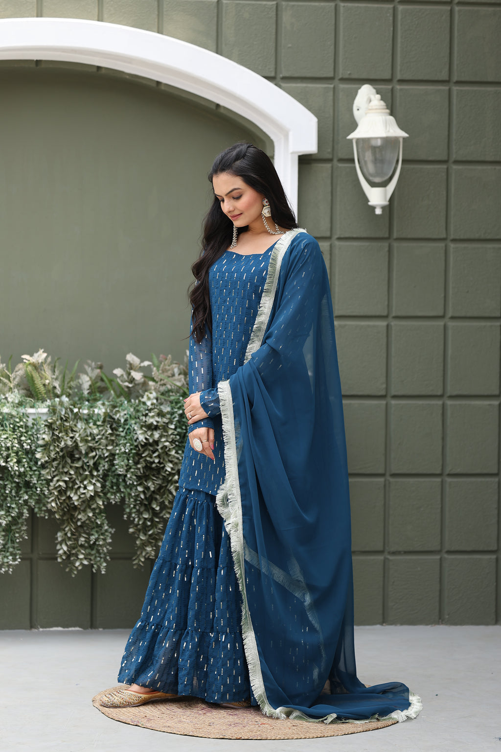 Woman in a blue traditional outfit standing against a green tiled wall with a decorative arch.