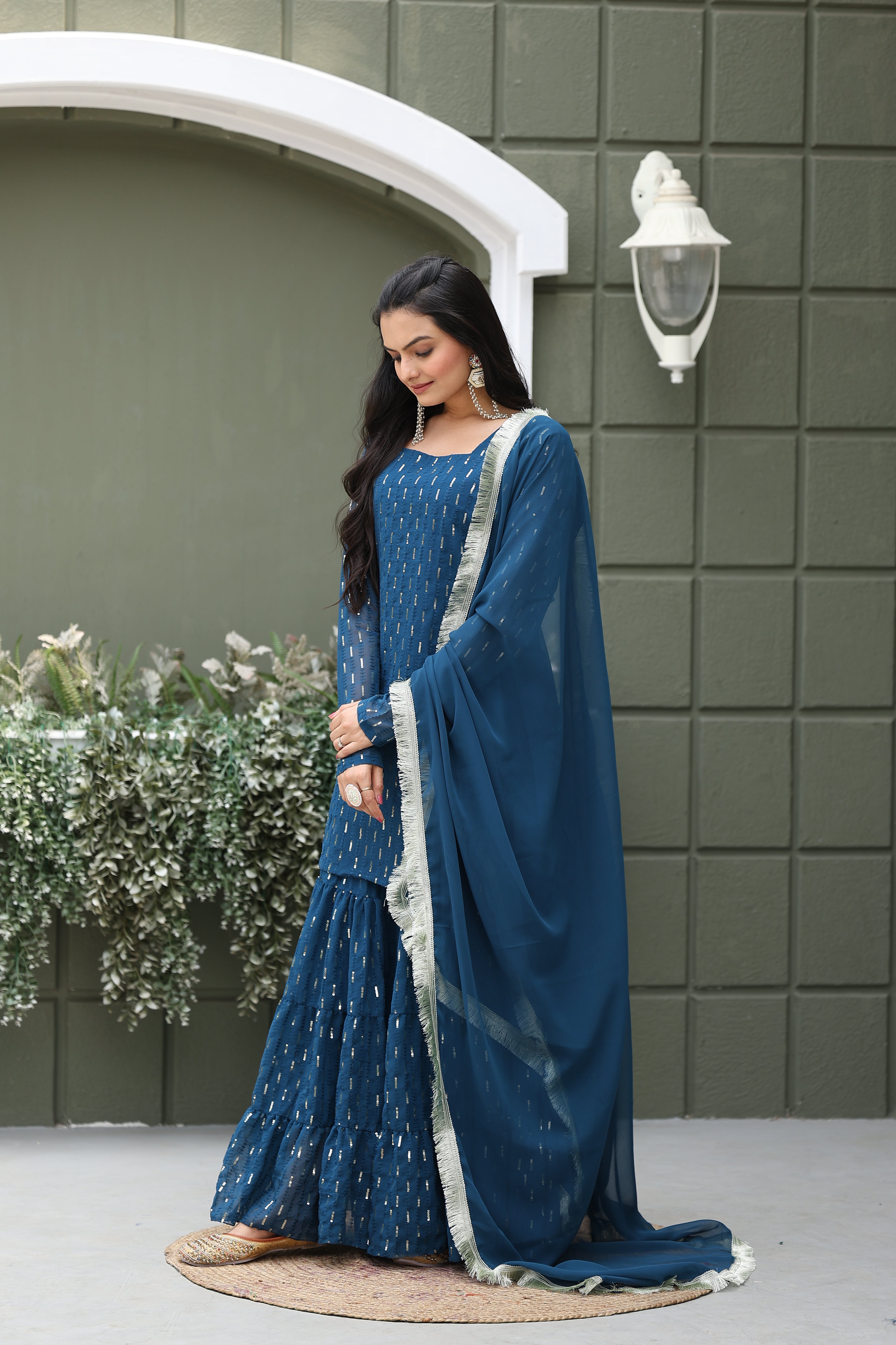 Woman in a blue traditional outfit standing against a green tiled wall with a decorative arch.