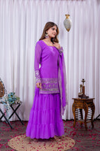 Woman in a purple traditional outfit standing against a white curtain background.