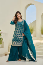 Woman in a teal traditional outfit with a patterned top and matching dupatta, standing in an outdoor setting.