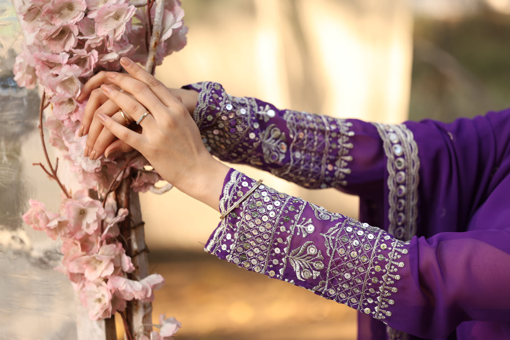 Person wearing a purple embroidered outfit holding pink flowers with a blurred natural background