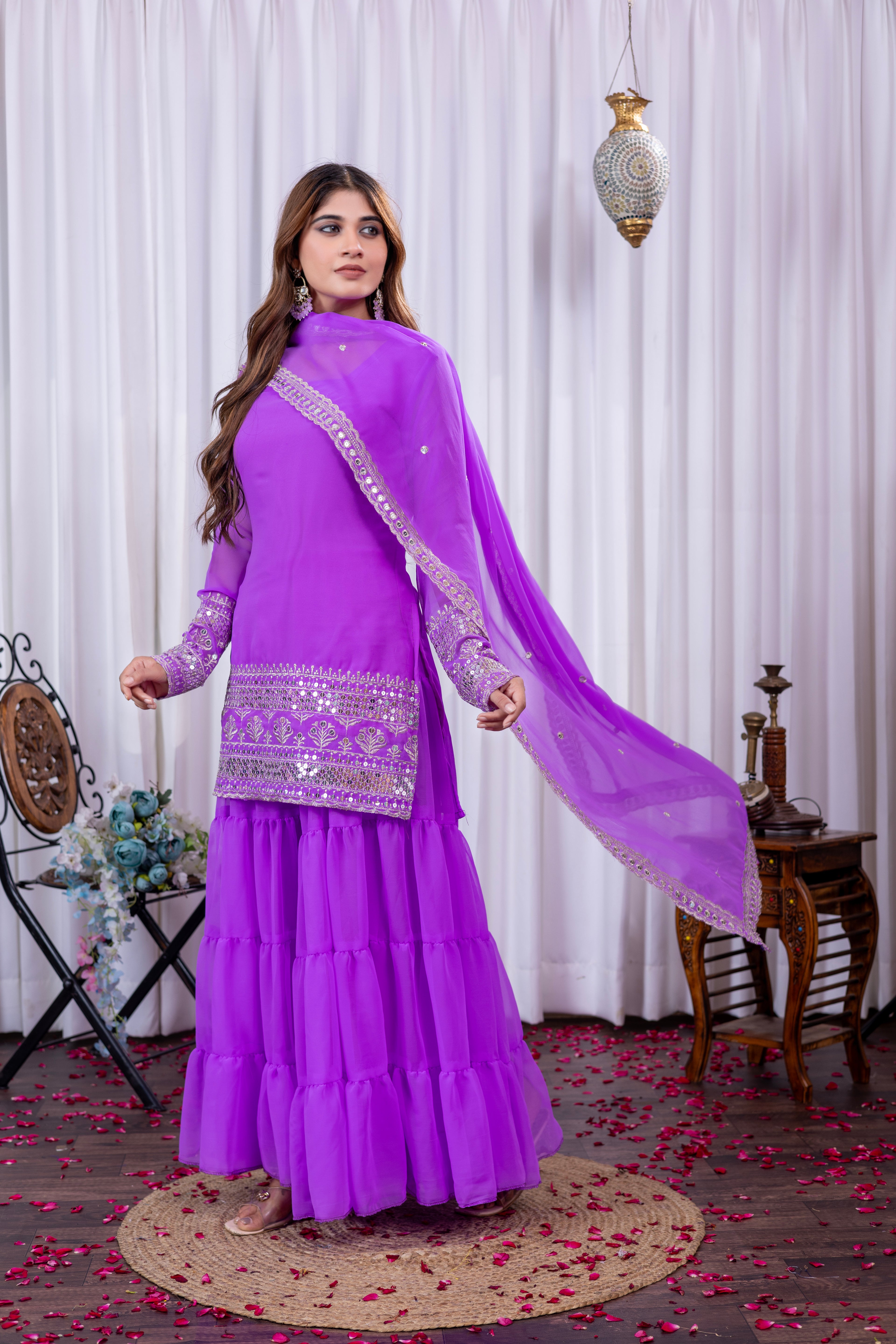 Woman in a purple traditional outfit with a white embroidered border, standing against a white curtain background.