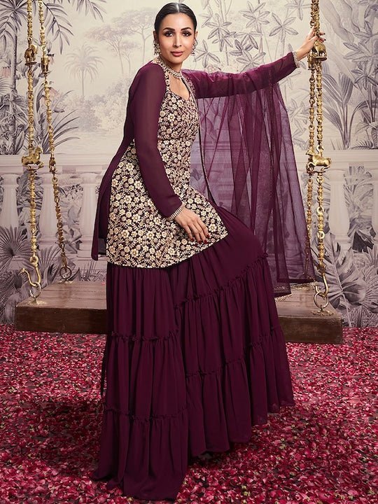 Woman in a traditional outfit with a purple top and sharara, standing on a swing with floral background.