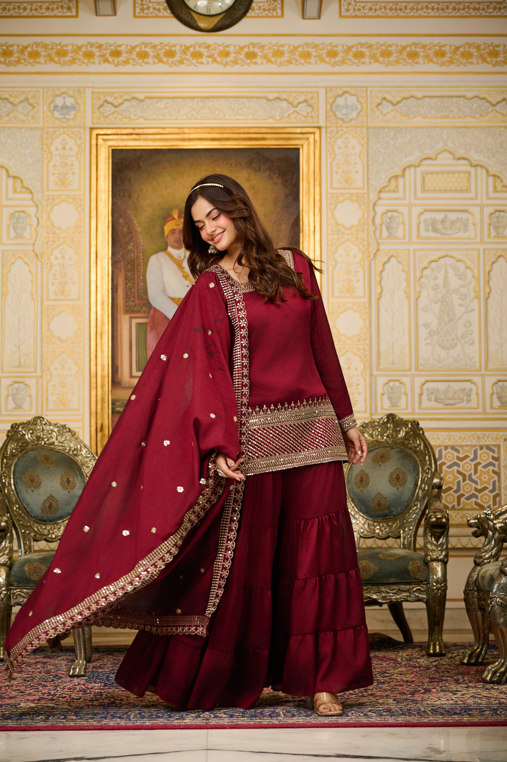 Woman in a red traditional outfit standing in an ornate room with decorative walls and furniture.