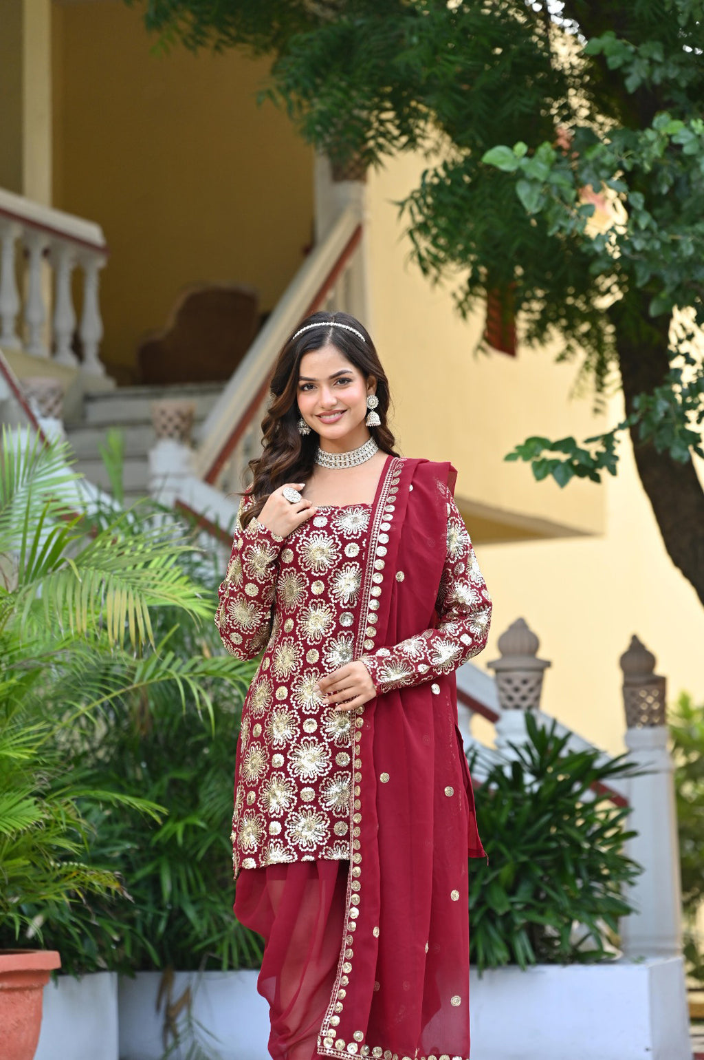 Woman in a red traditional outfit standing outdoors with greenery and a building in the background