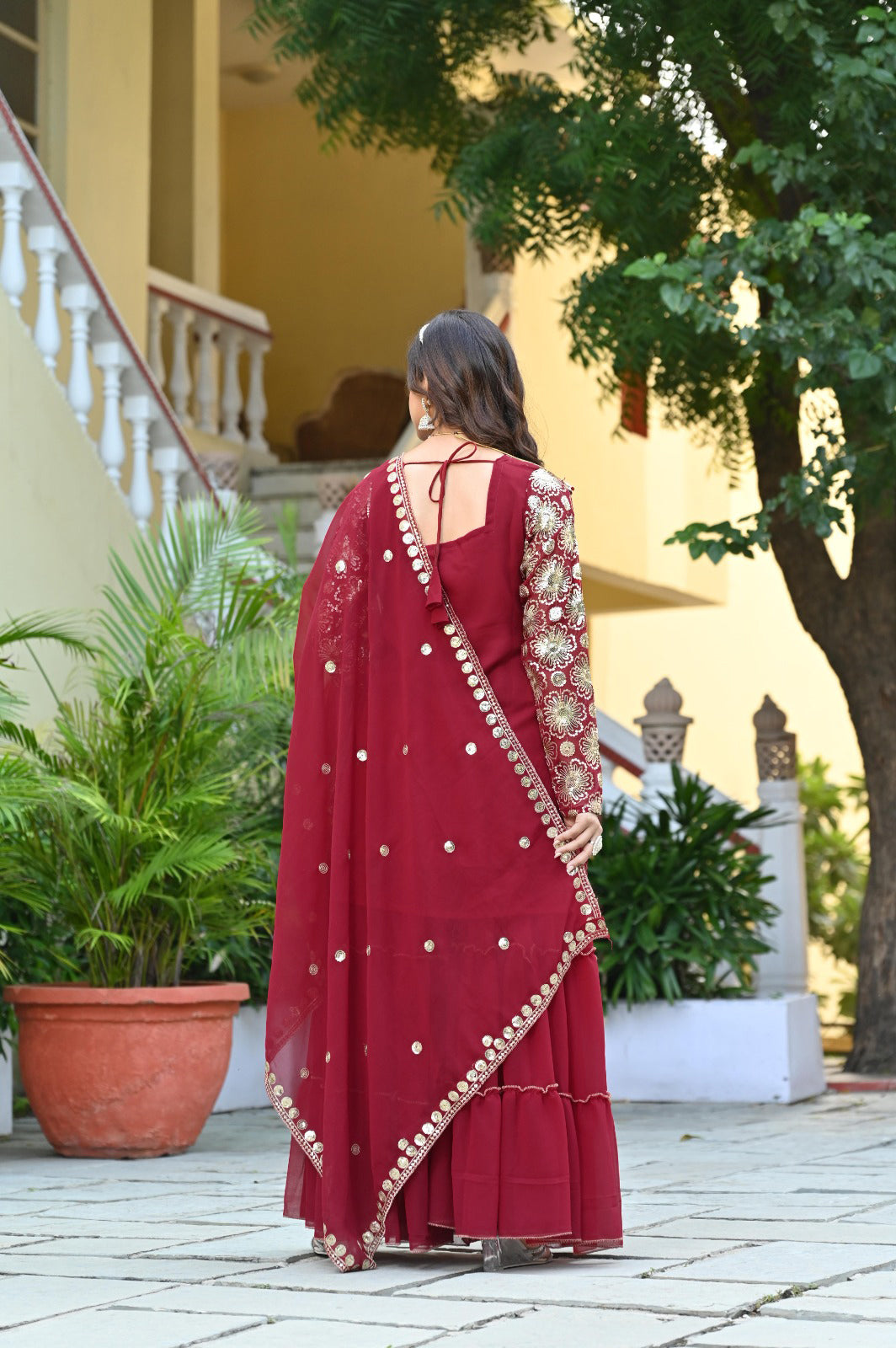 Woman in a red with gold details standing outdoors near a building and trees.
