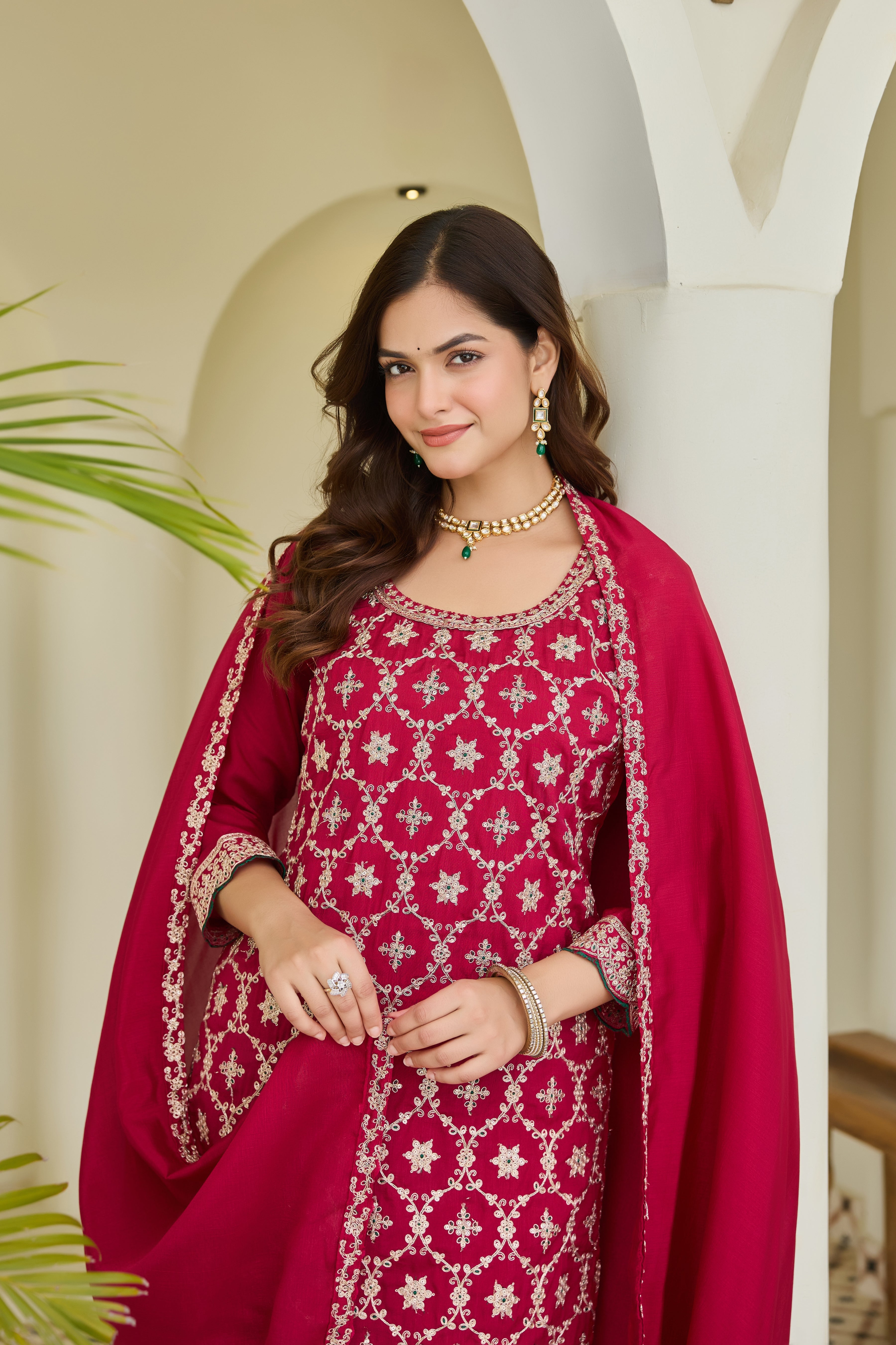 Woman in a red traditional outfit with intricate patterns, standing against a light-colored wall.
