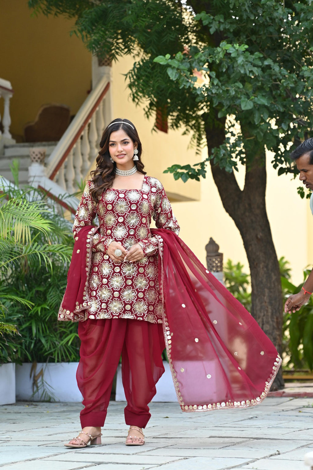 Woman in traditional maroon and gold outfit with a red dupatta outdoors.