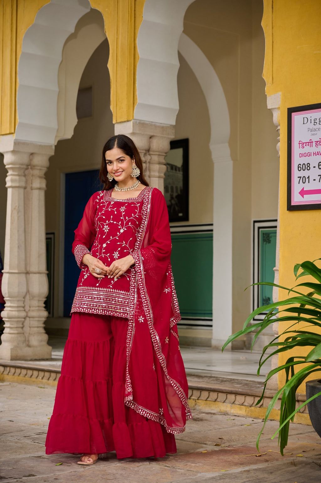 Woman in a red traditional outfit standing in front of a yellow building with arches.
