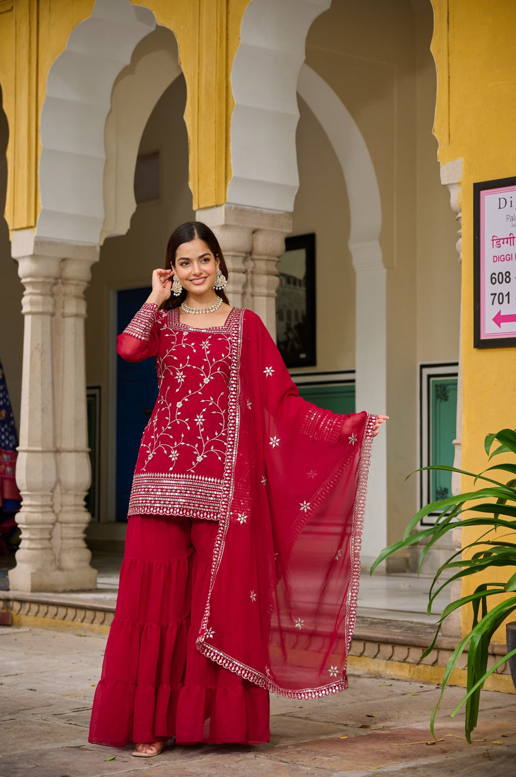 Woman in a red traditional outfit with white patterns standing in front of a yellow and white architectural structure.