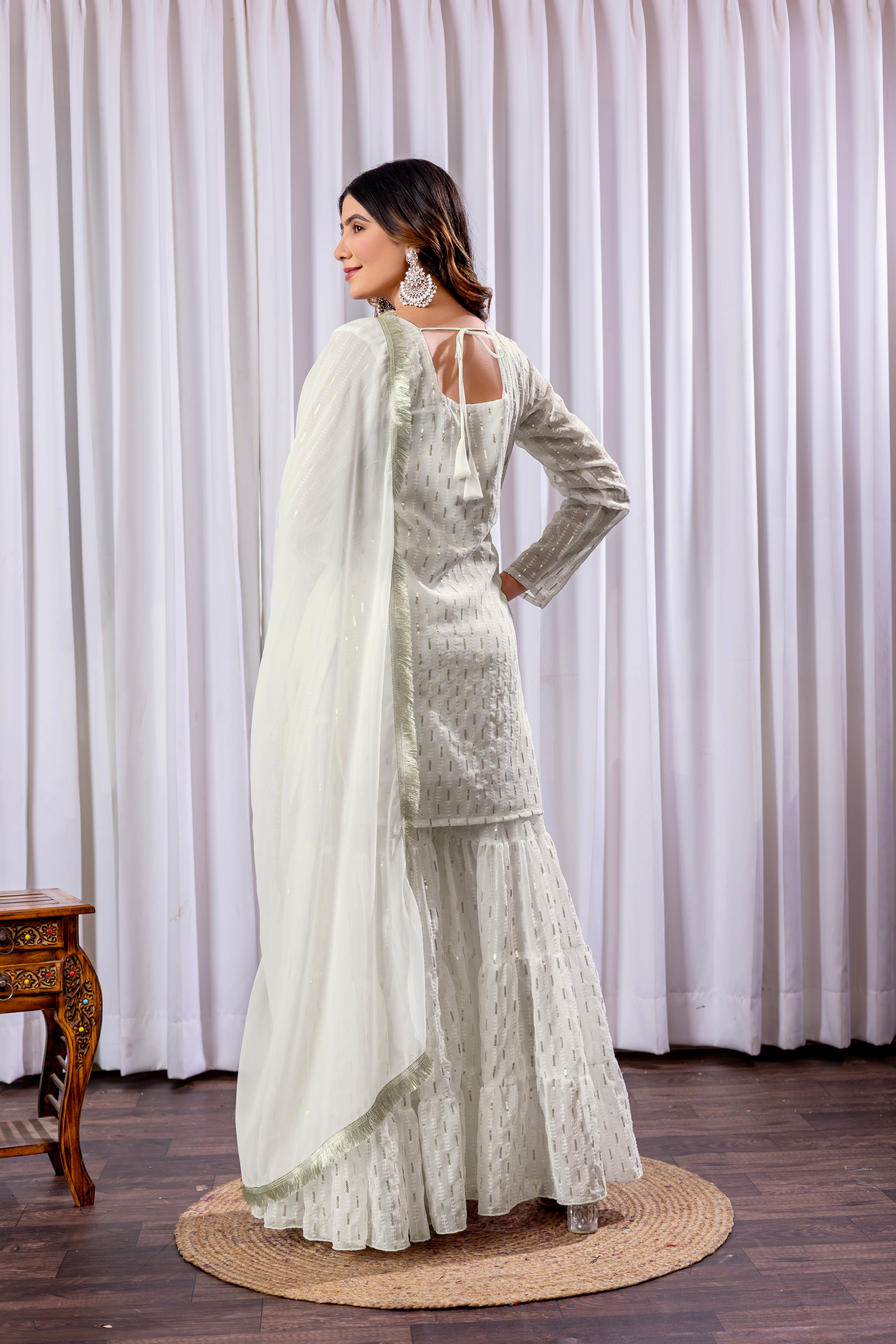 Woman in a white traditional outfit with a sheer dupatta against a white curtain background.